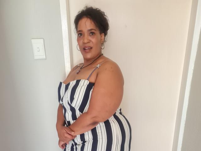 CaramelHugeAss is a curvy woman with dark curly hair, wearing a striped dress, standing with hands in front, smiling at the camera, showing her ass and small tits.