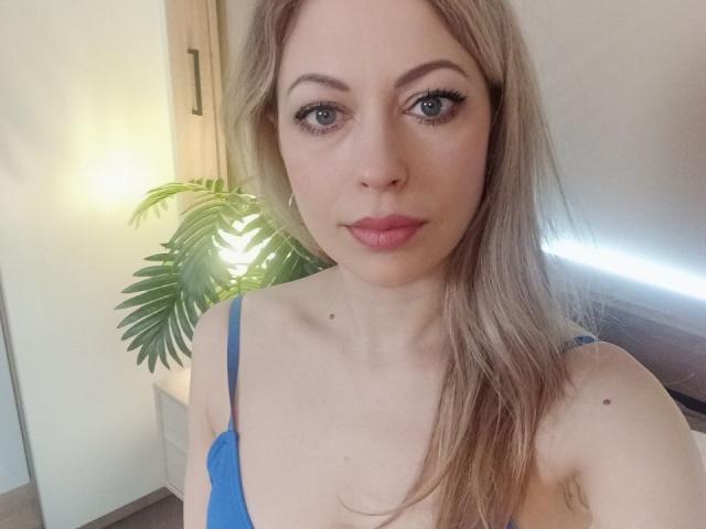 SweetLadyXX has light blue eyes, pink lips, and long blonde hair, wearing a blue lace bra, showing her small tits and a slight mole on her chest.