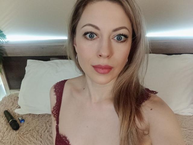 SweetLadyXX has blue eyes, light skin, and long blonde hair. She's wearing a red lace bra, with her tits slightly visible. A dildo is on the bed.