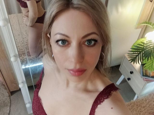 A selfie of SweetLadyXX with blonde hair, blue eyes, and pink lipstick, wearing a maroon lace top, showing her tits in a mirror reflection, with hoop earrings.