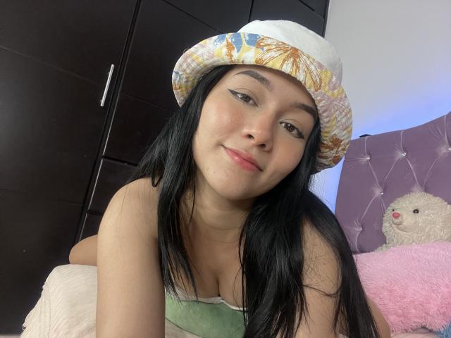 DeisyWest with long black hair, wearing a floral bucket hat, green top, and eyeliner, smiling at the camera, showing small tits, sitting on a bed with a pink pillow.