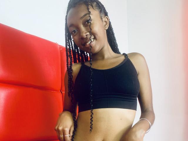 KarineHoney with dark skin, long braided hair, small black sports bra, showing her tits and midriff, red leather chair in background, smiling, wearing a silver bracelet.