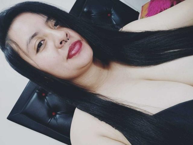 KatAndCharlotte has long black hair, red lipstick, and a nose ring, wearing a black strap top that shows her big tits. She's looking at the camera with a slight smile.