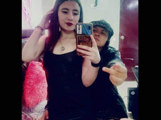 KatAndCharlotte with red lipstick, black dress, and necklace, holding phone, while Charlotte in black cap, pointing, takes selfie. KatAndCharlotte's tits visible under dress. Mirror reflection.