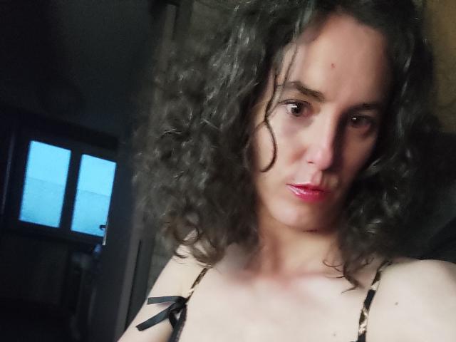 NinaAndGreg with curly dark hair, pink lips, and light skin, wearing a leopard-print top, showing small tits, looking serious, in a dimly lit room.