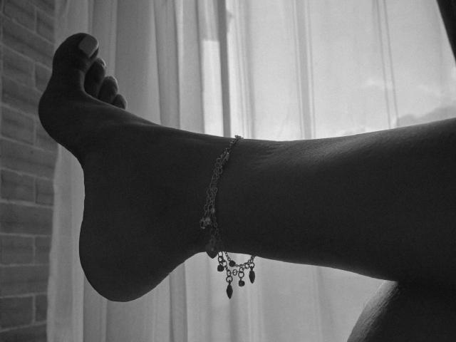 LunaMiranda's dark-skinned ankle with a beaded bracelet, her foot up, showing her toes against a bright window. Black-and-white, detailed, textured.