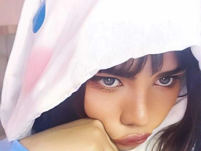 AnniieeBell with blue eyes, wearing a white hood with blue and pink dots, rests her chin on a yellow object, looking intensely at the camera. Her face is slightly flushed.