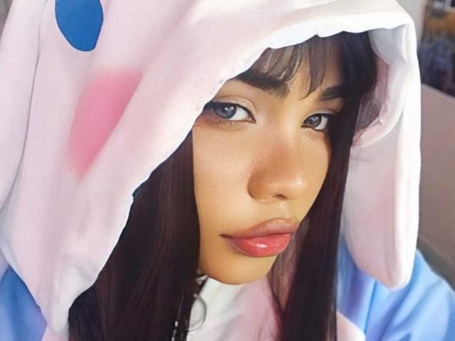 AnniieeBell with brown eyes and pink glossy lips, wearing a white and pink animal hood with blue and pink ears, has smooth brown skin and long dark hair.