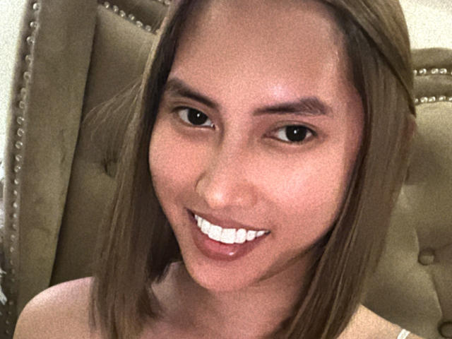BombshellPh has a cute smile with white teeth, brown eyes, and straight brown hair, wearing no clothes, sitting on a green, studded, upholstered chair.