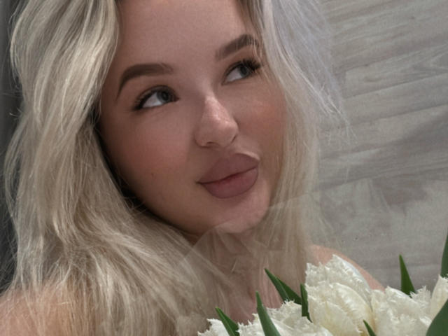 LollyTina with blonde hair, slightly wavy, holding white flowers, looking to the side with a subtle smile, soft makeup, visible nose, and green leafy stems in the bottom right.