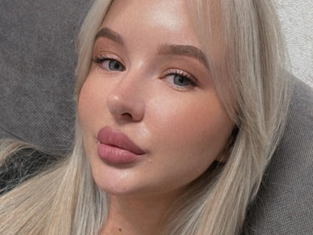 LollyTina has pale skin, blonde hair, and full pink lips with a slight sheen. She's making a pouty face with subtle makeup and blue eyes.