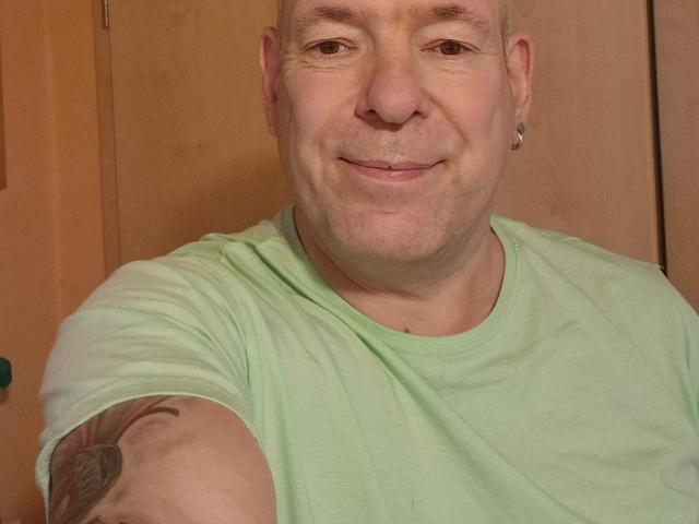 StrangerSixtySix has a bald head, light green t-shirt, tattoo on right arm, ear piercing, and a slight smile. Light skin, slightly chubby face. Wood door in background.