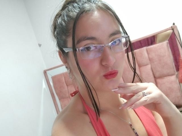 VenusSweetPalmer mit Brille, rotem Halstopp, rotem Lippenstift und einem Ring am Finger, die sich mit ihren feuchten, braunen Haaren das Kinn berührt. Brüste sichtbar, roter Hintergrund..