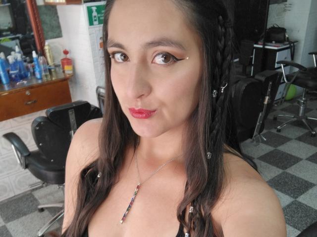 VenusSweetPalmer with dark braids, brown eyes, and red lipstick, wearing a black top with a necklace, showing her tits, in a salon with chairs and products.
