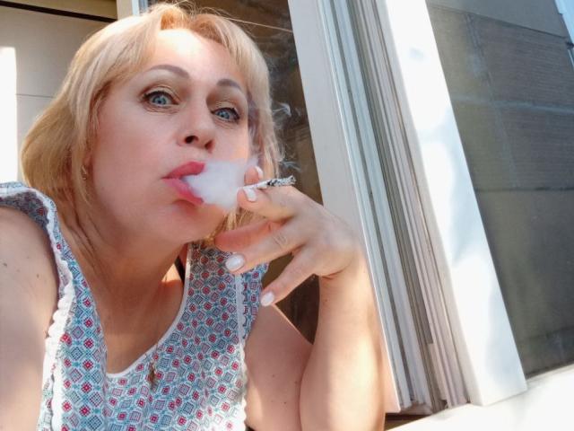 SusieNoreen mit blonden Haaren, blauen Augen und rosafarbenem Lippenstift, die an einer weißen Blase lutscht und ein oberteil mit Muster ohne Ärmel trägt, steht in der Nähe eines Fensters..