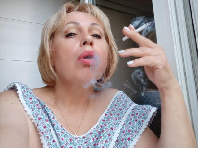SusieNoreen with short blonde hair, puffy pink lips, smoking a cigarette, blowing blue smoke, wearing a patterned dress, white nails, gold necklace, looking up.