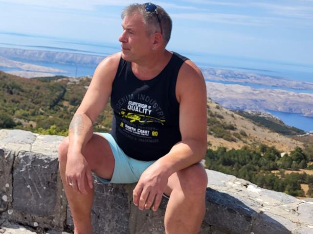 StrangerSixtySix, a chubby, middle-aged dude with a tan, wearing a black tank top and blue shorts, sits on a rock, looking off to the side with sunglasses on.