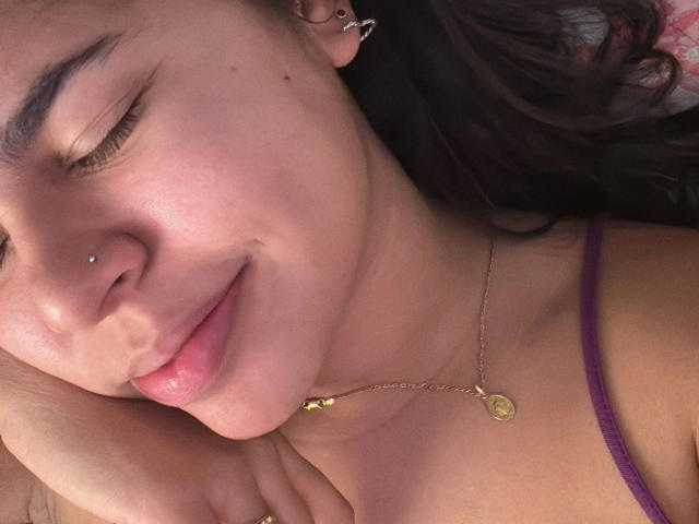 AnaMiss-hot with her eyes closed, nose pierced, gold necklace, and pink lips, smiling softly. She's wearing a purple strap, and her hand rests near her cheek.