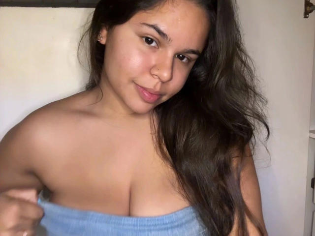 AnaMiss-hot with long dark hair, brown eyes, and a cute smile, holding a blue towel that barely covers her big, round tits. She's standing against a white wall.