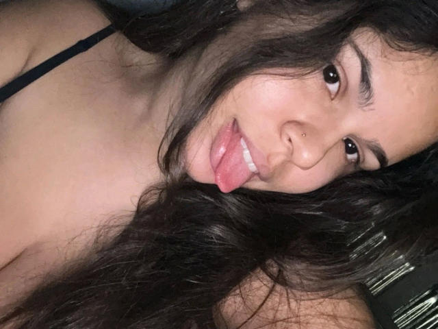 AnaMiss-hot with long dark hair, nose piercing, and pink tongue out, wearing a black spaghetti strap, has a cute, sexy, and playful expression.