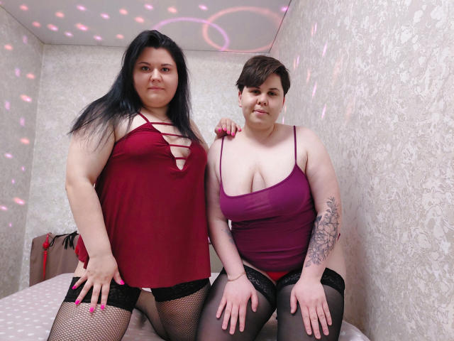 BeckyAndEllen with dark hair in red lace-up top, black fishnet stockings, standing beside BeckyAndEllen with short brown hair, purple top, black stockings, tattooed arm, and pink nipples.