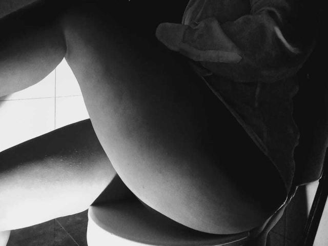 Black and white photo of JackelineRoset's ass, sitting on a chair, wearing a loose shirt, with legs slightly apart, showing smooth skin and subtle shadows.
