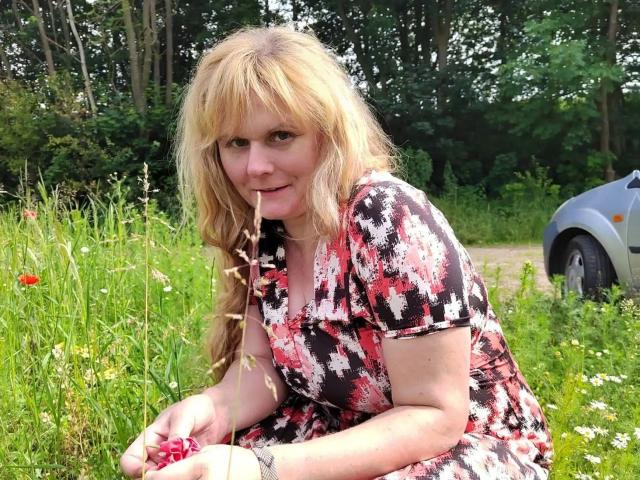 BeaLust-hot with long blonde hair, wearing a red and white floral dress, sucking a grass blade, sitting in a green meadow with wildflowers and a silver car.