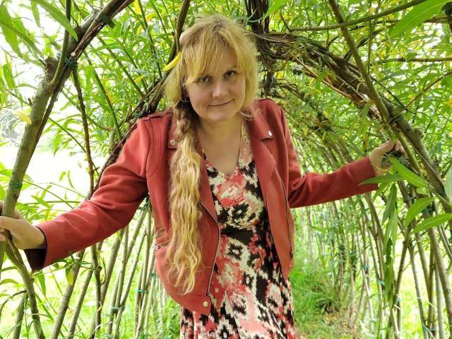 BeaLust-hot with blonde hair, wearing a red jacket and floral dress, standing in a bamboo grove, smiling slightly, holding bamboo sticks on both sides.