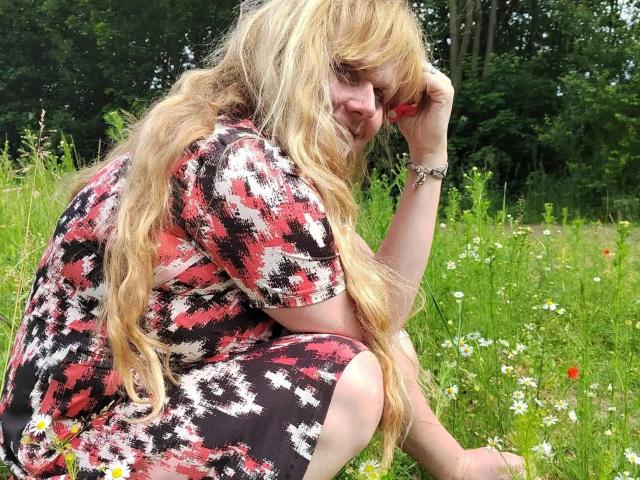 BeaLust-hot with long, messy blonde hair, wearing a red and black patterned dress, sits in a grassy field, touching her forehead with a slight, sad smile.