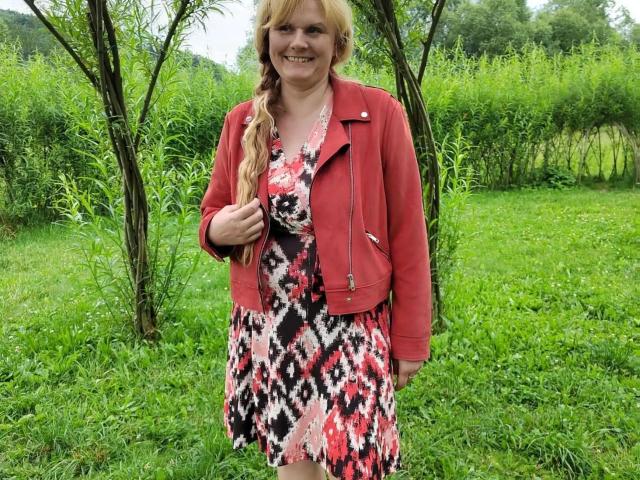 BeaLust-hot with blonde hair in a braid, red jacket, and patterned dress, standing on green grass, holding her hair, smiling, with trees in the background.