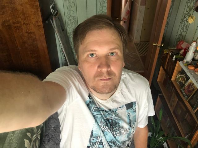 Selfie of SteelAlex with pale skin, blue eyes, light brown hair, white t-shirt, slightly chubby, taking photo with extended arm, in a cluttered room with wooden furniture.