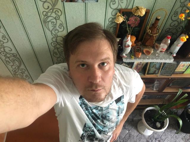 A selfie of SteelAlex with short brown hair, wearing a white t-shirt and green patterned suspenders, standing in a room with floral wallpaper and a wooden shelf.