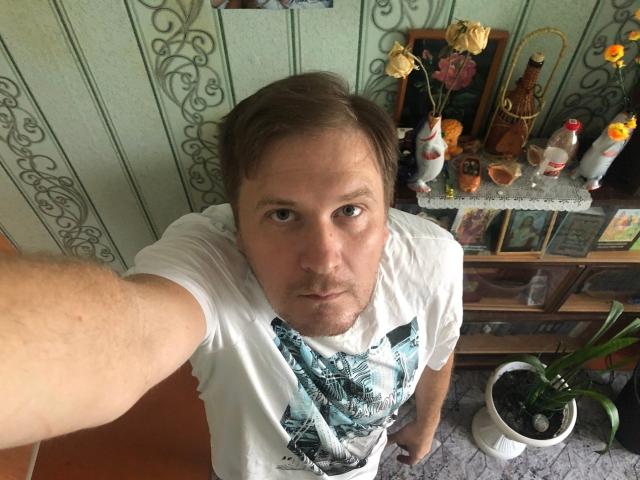 A selfie of SteelAlex with short brown hair, a white T-shirt, and a blue graphic, looking up with a serious expression, standing in a room with ornate green wallpaper.