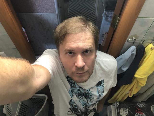 SteelAlex with short, blonde hair, white t-shirt, and slight beard, taking a selfie in a bathroom, looking up at the camera with a serious expression.