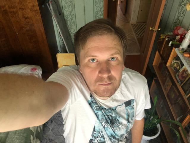 SteelAlex with light brown hair, blue eyes, and a beard, wearing a white t-shirt with a city print, taking a selfie, showing a slightly confused or tired expression.