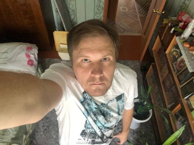 A selfie of SteelAlex, a white man with short brown hair and blue eyes, wearing a white graphic t-shirt, looking up with a serious expression. His hand is holding the camera.