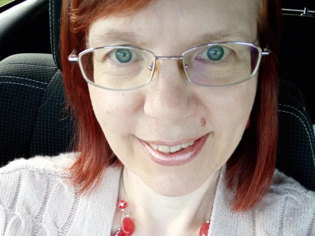 JuicyLady69 with red hair, glasses, blue eyes, smiling, light skin, small nose bole, pink lipstick, white knit top, red bead necklace, sitting in a car.