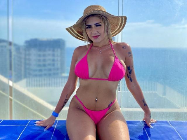 SusiAndMat with big tits in a pink bikini, wearing a straw hat, sitting on blue tiles, tattoos on arms, blue bracelet, belly button piercing, ocean and city in background.