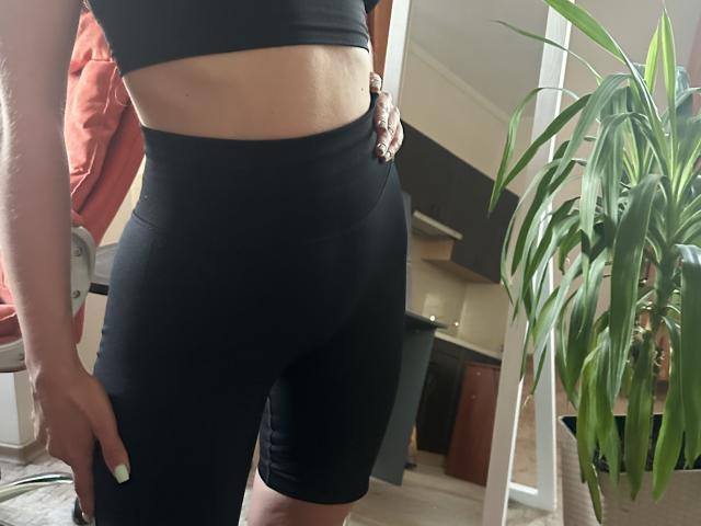 Stefanni's black booty shorts hug her tight, round ass, and her hand rests on her hip, showing a bit of her midriff, with a plant to the right.