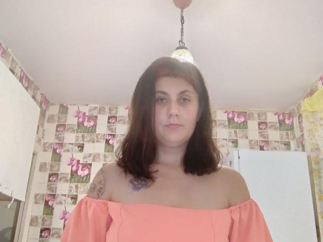 DianeCordy with shoulder-length brown hair, wearing an off-shoulder pink top, has a small tattoo on her right shoulder and a neutral expression, standing under a ceiling light.