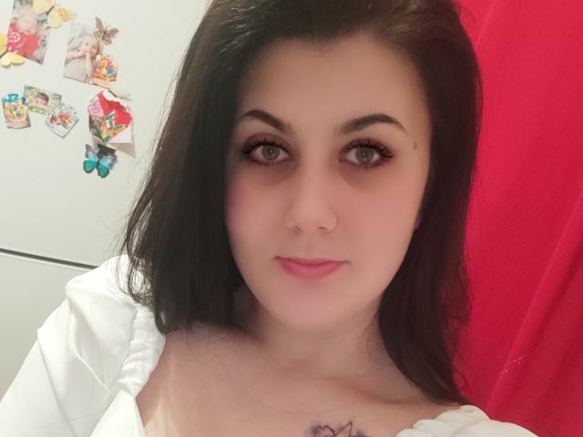 DianeCordy with pale skin, dark brown hair, and pink lips, wearing a white top, showing a small purple tattoo below her tits, and looking at the camera.
