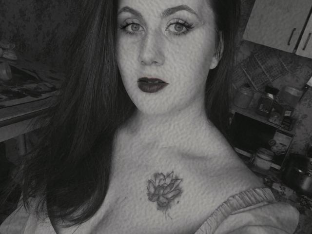 Diane Cordy, com cabelos longos e escuros, usando batom escuro, mostrando uma tatuagem de uma flor no peito, em uma foto em tons de cinza, com os seios levemente visíveis sob uma camisa folgada..