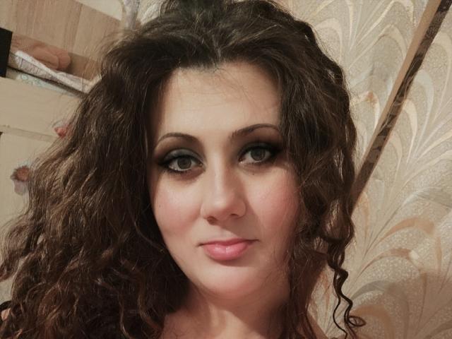 Diane Cordy mit lockigem, braunem Haar, grünen Augen und rosafarbenen Lippen, die in die Kamera blickt und dabei ein leichtes Lächeln zeigt, mit dunklem Eyeliner und rosafarbenem Lippenstift..