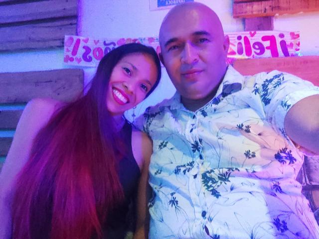 KratosYHotties with long red hair and big smile, wearing black top, next to bald KratosYHotties in white tropical shirt, both sitting, purple and pink light.