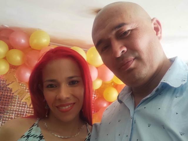 KratosYHotties has bright red hair, small tits, and a smile, wearing a patterned top. Bald KratosYHotties in a blue shirt stands next to her, smiling.