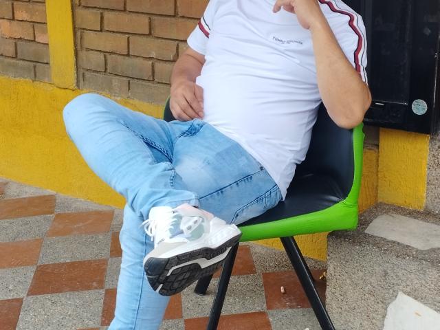 KratosYHotties with a chubby belly, white shirt, blue jeans, and white sneakers with green accents, sitting on a green chair, leg crossed, hand near face.