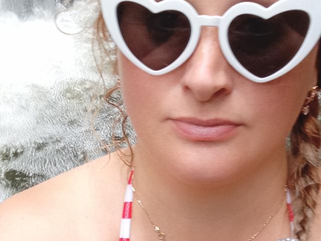 KelliRedhaer trägt weiße, herzförmige Sonnenbrillen, lockiges blondes Haar, rosafarbene Lippen und ein rotes und weißes Halstopp. Sie trägt eine goldene Halskette. Ihr Gesicht ist leicht gebräunt..
