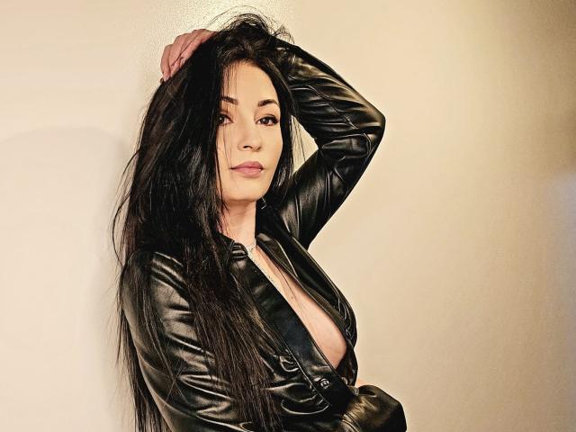 Coryna, avec de longs cheveux noirs, portant une veste en cuir noir, dévoilant sa poitrine, une main dans les cheveux, regardant la caméra, lumière douce, lèvres légèrement entrouvertes, expression confiante..