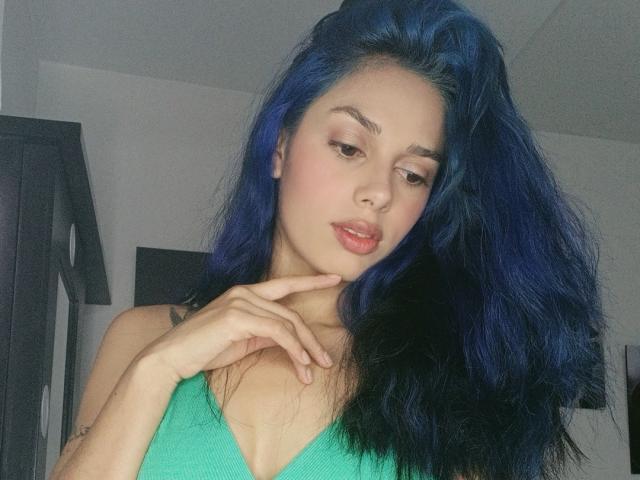 AnaisHilton with blue hair, green top, and pink lipstick, looking down with her finger on her chin, showing her tits and a tattoo on her left arm.