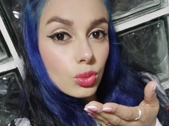 AnaisHilton with blue hair, pink lipstick, and winged eyeliner, pouting lips, holding hand with pink nails, ring on finger, brown eyes, and slightly parted eyebrows in a selfie.
